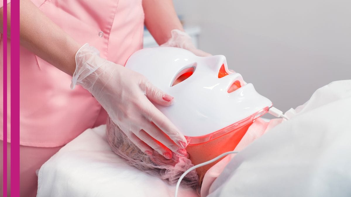 A red light therapy mask is placed on a client's face. The esthetician has discussed red therapy benefits.